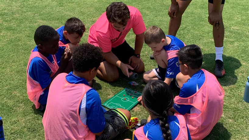Youth soccer players kneel around a board where a coach is pointing out alignment instructions