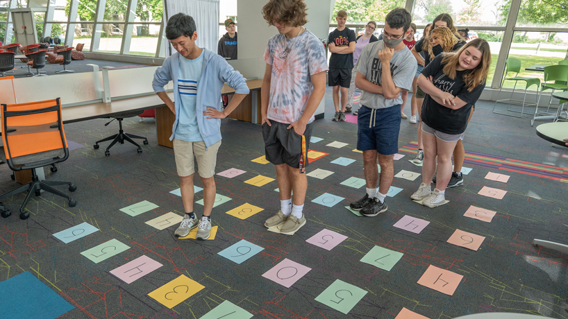 Students stand and look at numbered pieces of paper laid out on the floor as they contemplate their next move in solving a puzzle