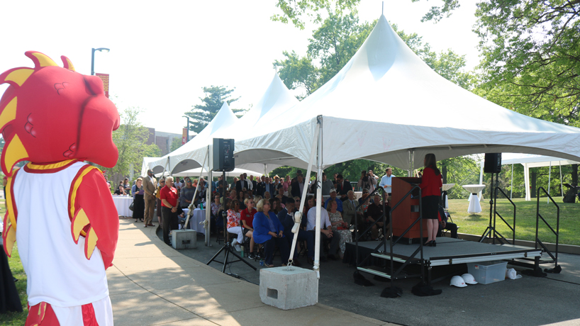A crowd gathers under the shade of a tent to listen as Vice Chancellor Lisa Capone speaks during a groundbreaking ceremony for the Richter Family Welcome and Alumni Center