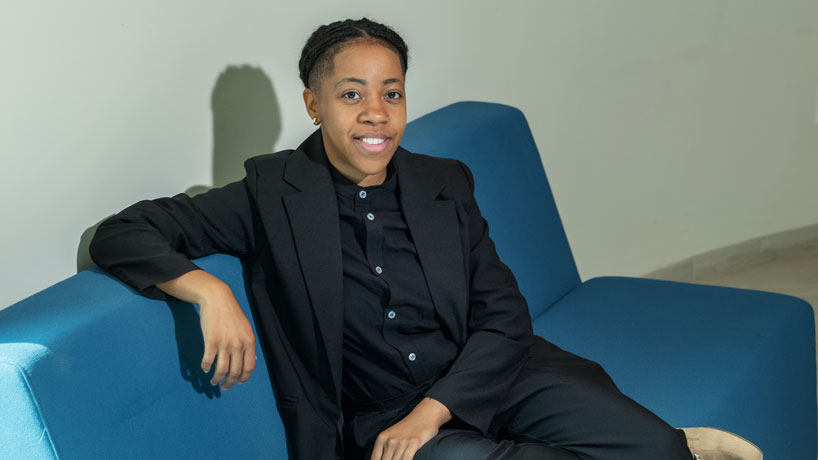 Young Black woman sits on couch in a black suit looking up at the camera.