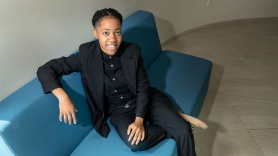 Young Black woman sits on blue couch in black suit looking up at the camera.