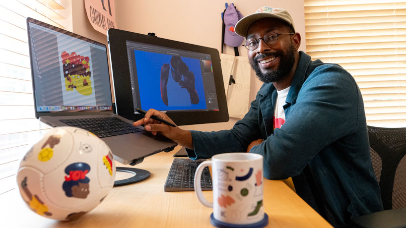 Young Black man, casually dressed, wearing glasses, sits in front of a computer screen next to a soccer ball that features different characters.