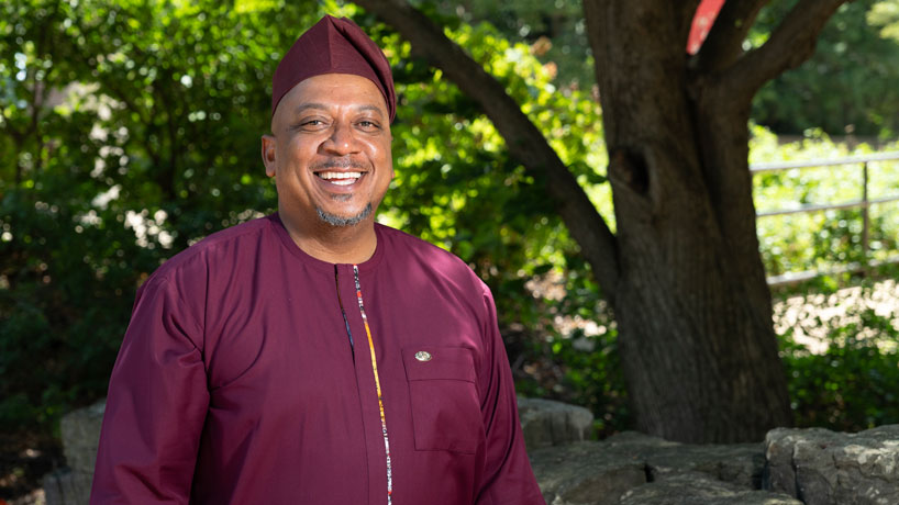 Black man wearing African clothing stands outside in front of trees smiling.