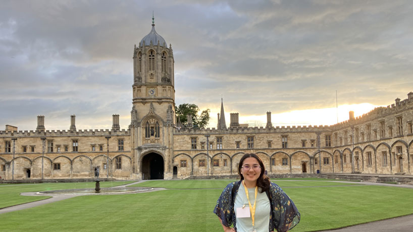 Young Asian woman stands in front of a massive castle-like structure, Tom Tower in London.