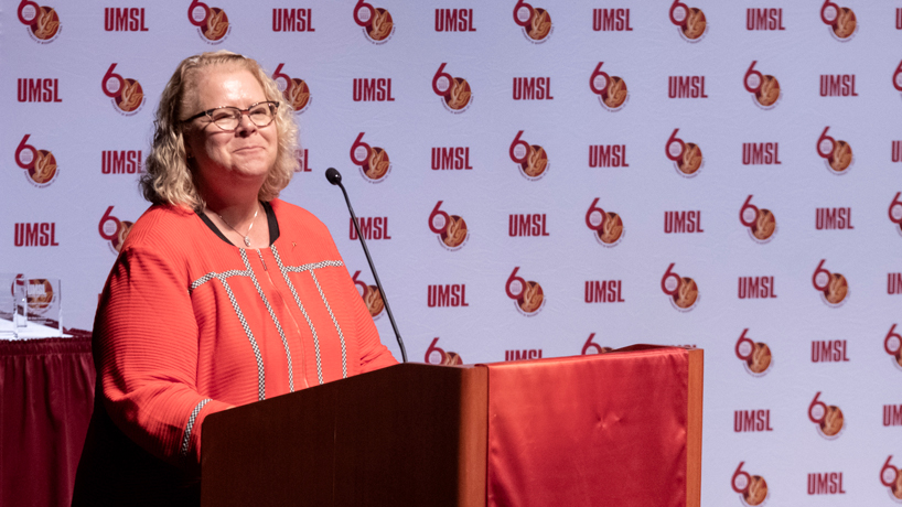 Chancellor Kristin Sobolik delivering the State of the University Address while standing behind a lectern with a backdrop including the UMSL and 60th anniversary logos in the background