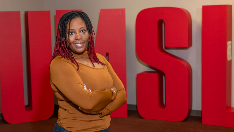 Black woman wearing brown top stands in front of an UMSL logo background.