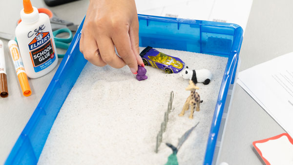 A women's hand reachs into a sand tray with small children's toys
