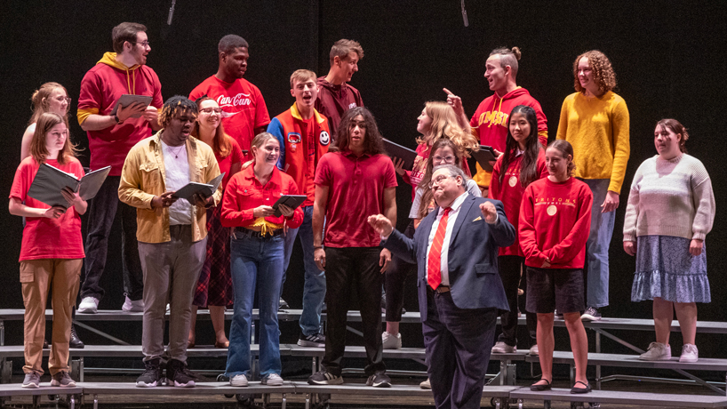 Students wearing UMSL sweatshirts and T-shirts stand on risers and sing on stage under the direction of Associate Professor Jim Henry