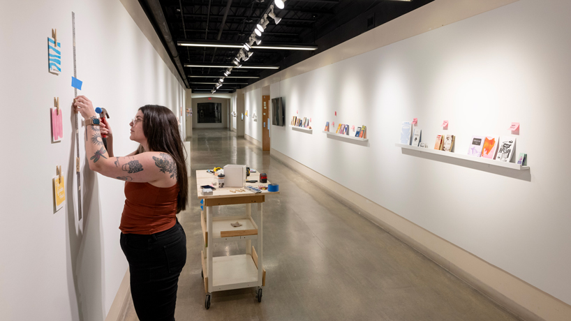 Taylor Timmerberg, a University of Missouri–St. Louis graduate and now a senior research lab/print technician in the Department of Art and Design, works to install the “Printed with Purpose” Zine Show + Book & Risograph Art last week at Gallery 210@FAB