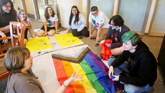Several people are sitting on the floor in a room around around a big Pride flag drawing figures on it.