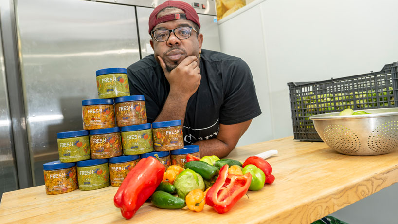 Jordan Franks leans over a counter with a pyramid of Taste of Jack's salsa and an array of peppers and tomatoes