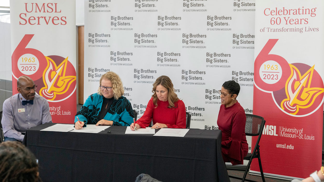 University of Missouri–St. Louis Chancellor Kristin Sobolik and Big Brothers Big Sisters of Eastern Missouri CEO Kristen Slaughter sign a memorandum of understanding in front of UMSL and BBBSEMO banners as Derrick Freeman and Eboni Buckles look on