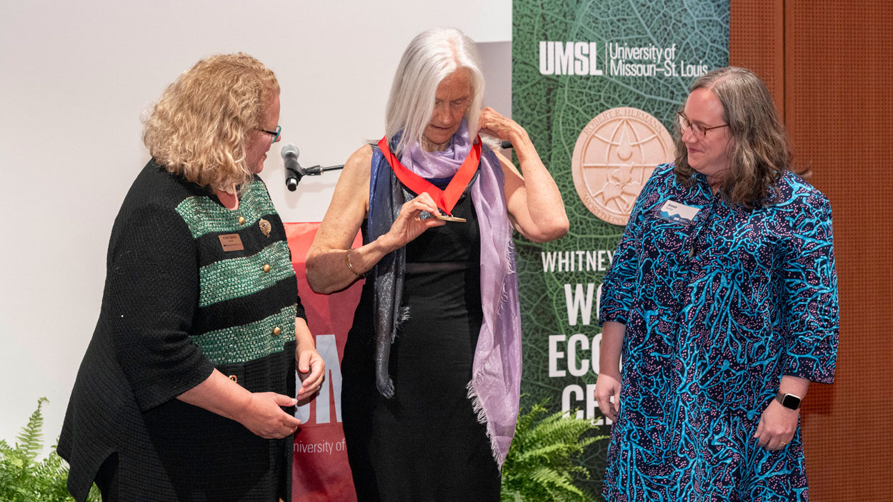 Julie Packard looks at the Robert R. Hermann World Ecology Award medal after accepting it from UMSL Chancellor Kristin Sobolik as Whitney R. Harris Center Interim co-Director Aimee Dunlap looks on