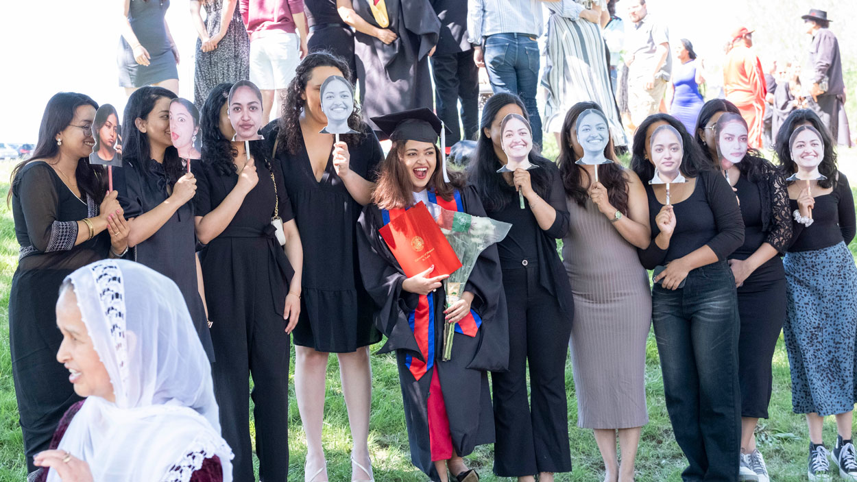 A graduate takes photos with friends and family members holding up masks in front of their faces after commencement
