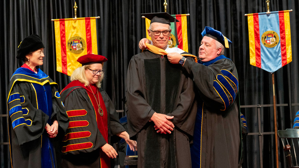 Provost Steven J. Berberich places a hood around retired Ameren Executive Chairman Warner Baxter while presenting him with an honorary doctorate as College of Business Administration Dean Shu Schiller and Chancellor Kristin Sobolik look on