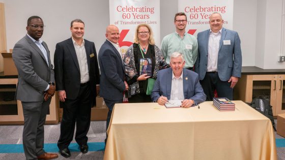 Mike Parson sits at a table signing books while surrounded by Reggie Hill, Reda Amer, Robert Sharp, Kristin Sobolik, Blake Sutter and Matt Prsha