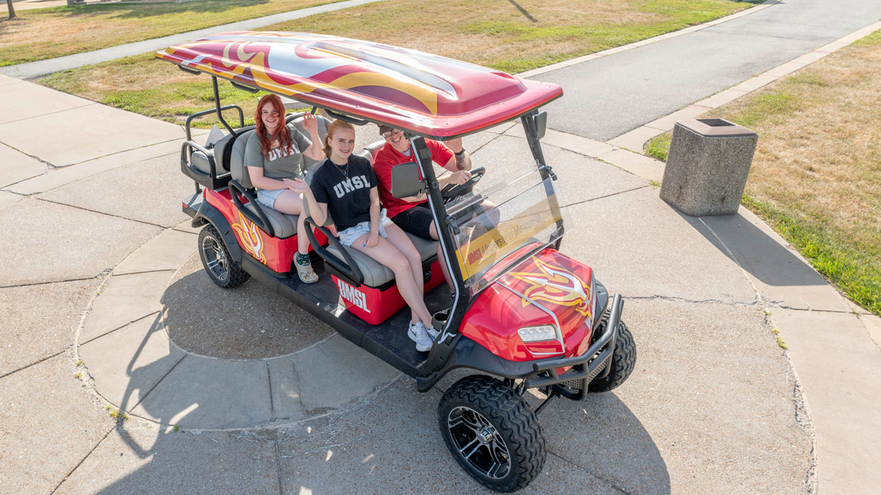Triton Leaders Allison Lendman, Ashley Schauwecker and Cole McWilliams seated in the newly wrapped, UMSL-branded red golf cart outside the Millennium Student Center
