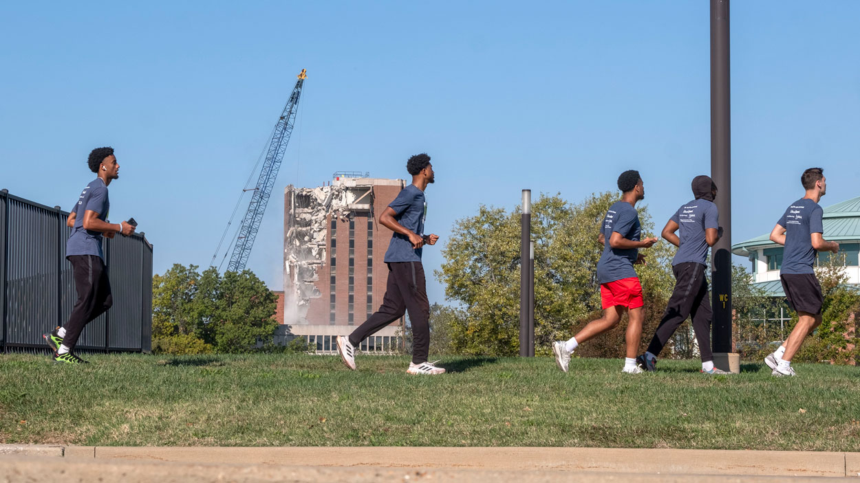 A crane takes a wrecking ball to the Social Sciences and Business Building Tower Saturday morning on North Campus at the University of Missouri–St. Louis as a group of students runs in the foreground along Grobman Drive while taking part in the fourth annual UMSL Alumni Association 5K Run/Walk.