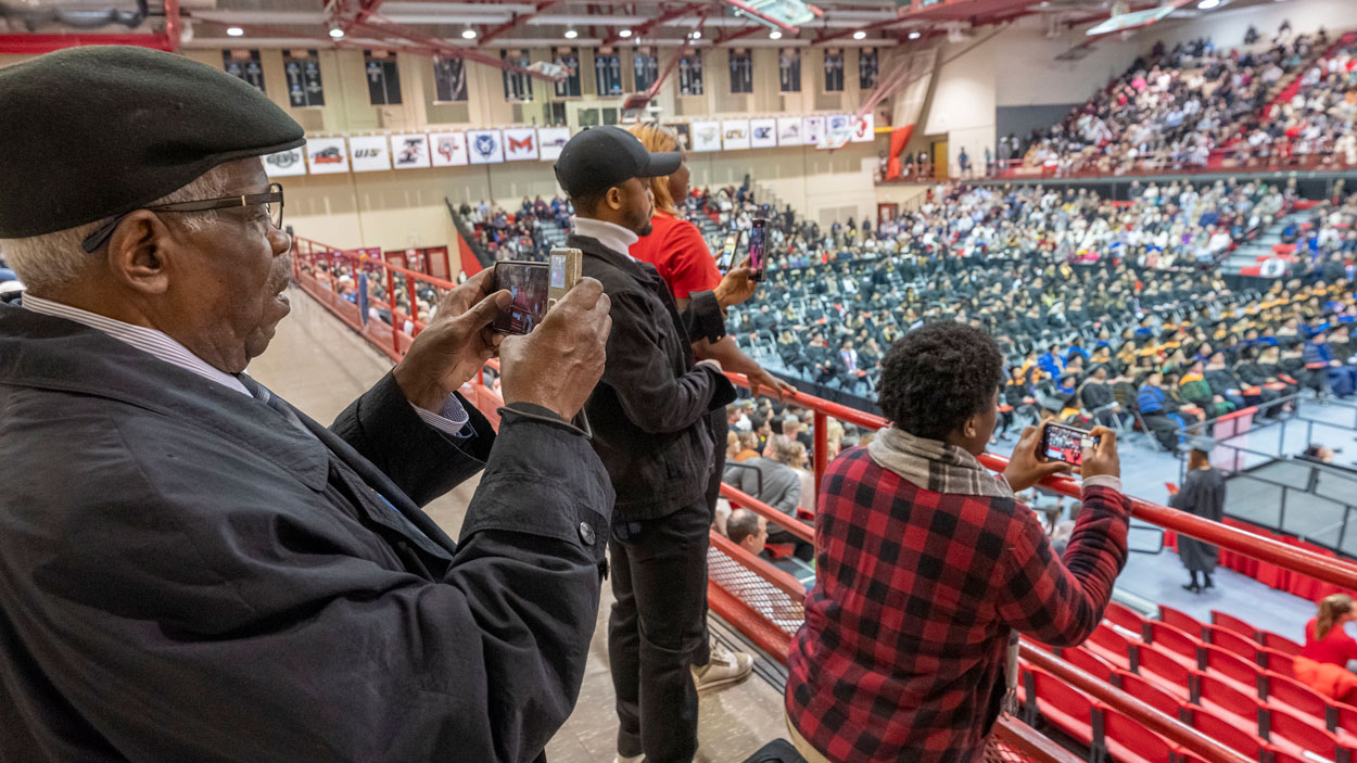 Family members stand to take pictures of the commencement stage from their seats in upper section of the Mark Twain Athletic Center.