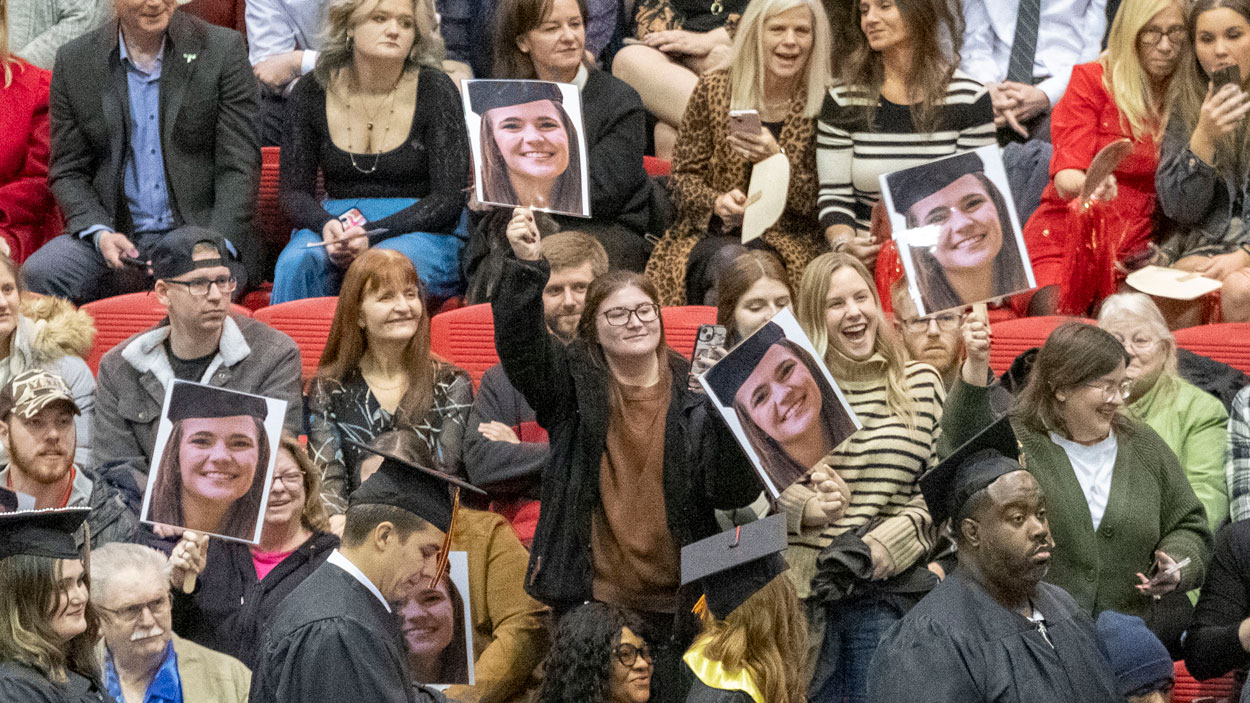 Friends and family members hold up pictures of their graduate as she walks across the commencement stage