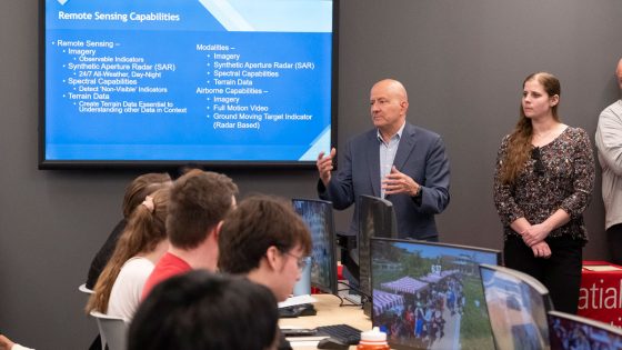 Retired Vice Admiral Robert Sharp speaks to students in the Geospatial Advanced Technology Lab as Samantha Dobson stands and listens