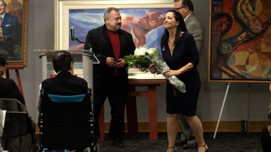 Bess Fitzmaurice receives flowers from the Hellenic Spirit Foundation as artist Rip Kastaris looks on from behind the lectern.