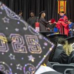 A mortarboard decorated with 2025 is in the foreground as a graduate walks across the commencement stage