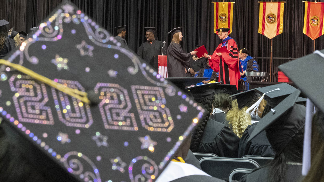 A mortarboard decorated with 2025 is in the foreground as a graduate walks across the commencement stage