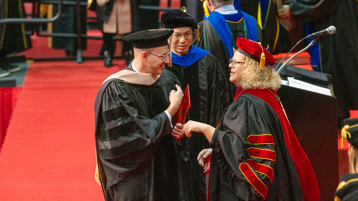 Ed G. Smith College of Business Doctor of Business Administration graduate, Karl Guenther, Yiuman Tsu, PhD and Chancellor Kristin Sobolik share a moment on stage after Guenther was hooded by Tsu for his DBA