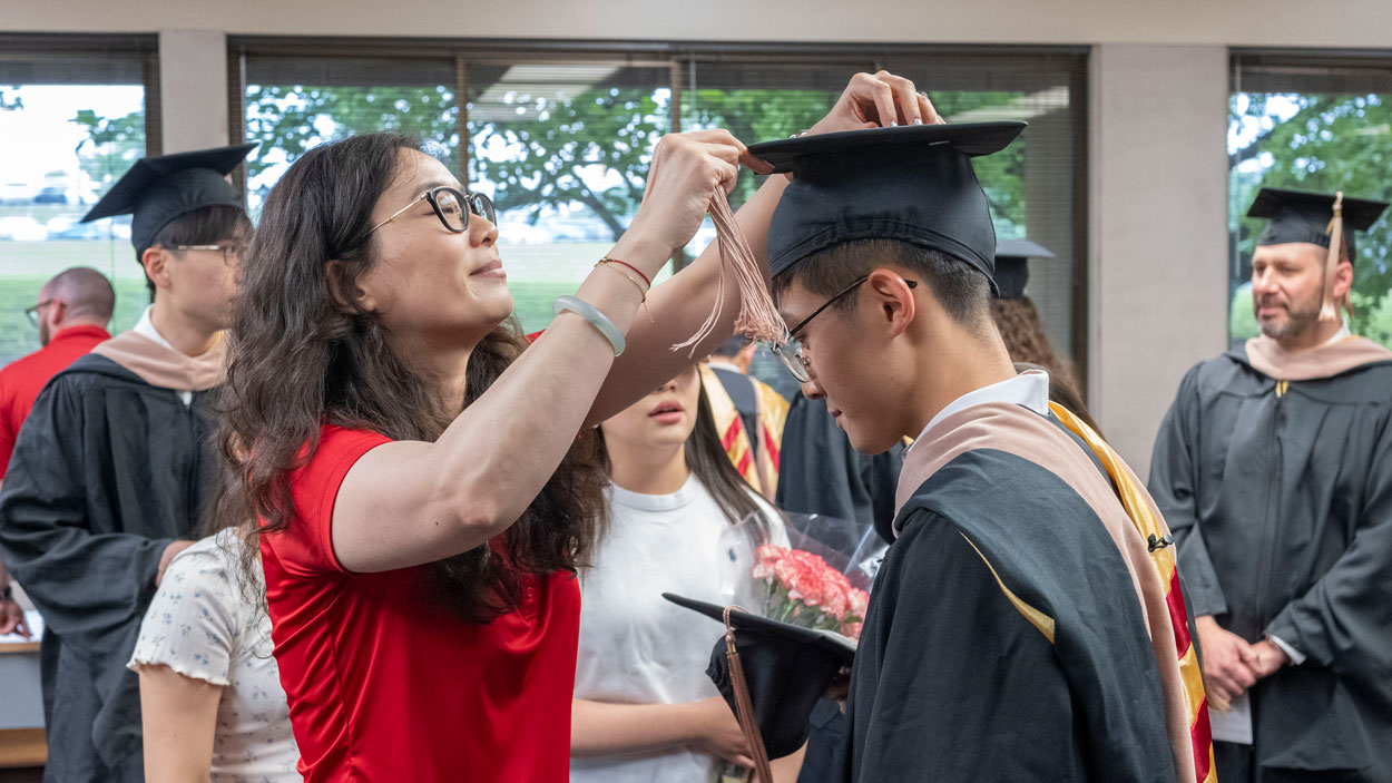 Yuan Chen, director of business student success, helps a graduate student in the College of Business with their tassle before the start of commencement. 