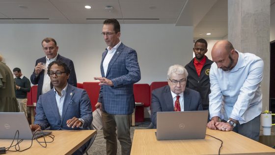 Two men sitting at laptop computers getting instruction from people standing next to them